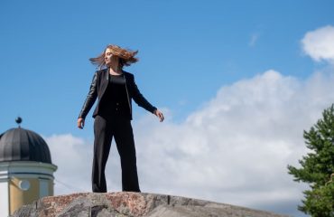 Young woman posing confidently on a rock in a Helsinki park, with a clear blue sky and European architecture in the background.